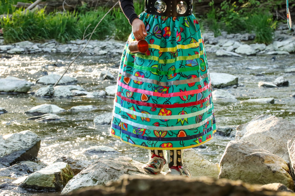a woman stands in a river with a long multicoloured striped skirt of ribbons